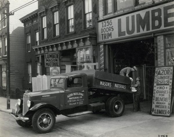 Workers unloading bags of "plaster of paris" from the back of an International dump truck owned by Azor Building Material Company. Signs on the building and truck advertise mason's materials and lumber.