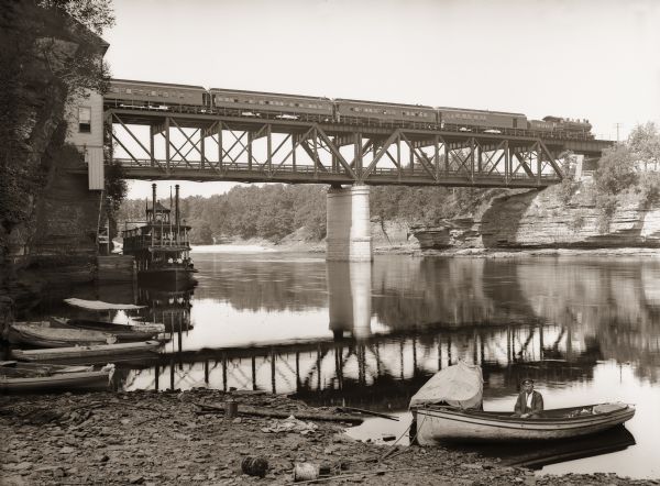 Chicago, Milwaukee and St. Paul passenger train on Kilbourn railroad bridge. "Apollo No. 1" steamboat below bridge, with musicians in bow. Various launch boats at shore. Man sitting in launch boat "Eva" in the foreground.