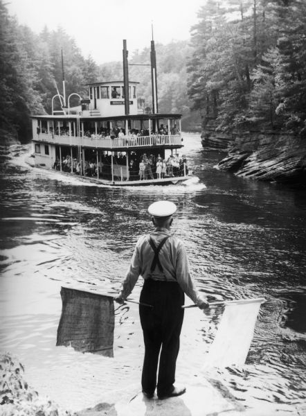 Rear view of flagman standing at the narrows in the Wisconsin River as a steamboat approaches.