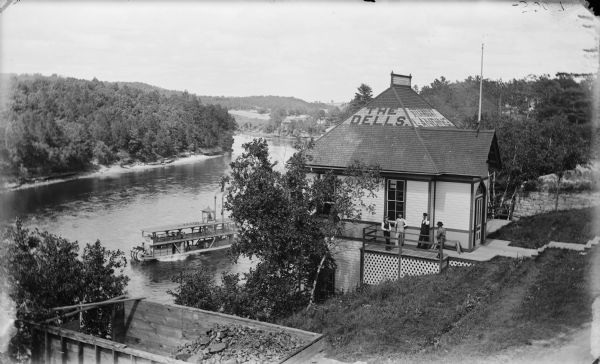 Elevated view of the Dells boat landing building, with people standing on the balcony. There is a steamboat near the dock on the river below.
