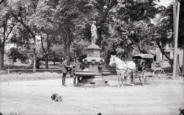 Two men are standing by the fountain on Broadway, which is topped by a statue. A horse and carriage are parked next to the fountain, and a dog is relaxing on the road in the foreground.