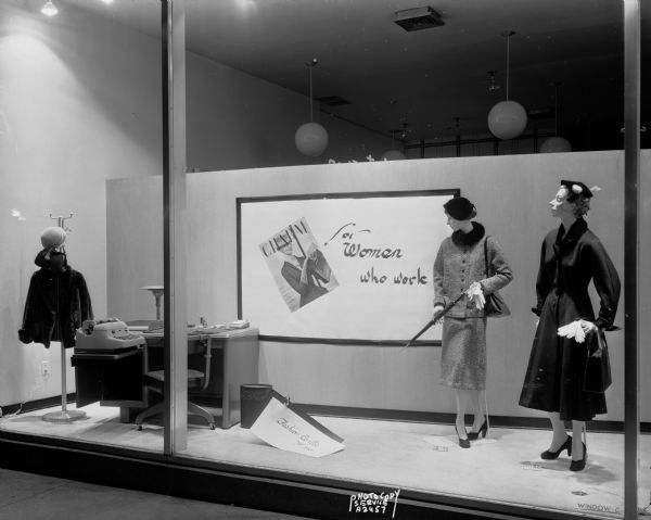 Manchester window display featuring outdoor clothing for "Women Who Work," with two mannequins, an office desk and a clothing stand with a jacket, hat and purse, window C.