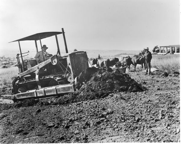 Man moving earth in South Africa with an International TD-14A crawler tractor equipped with a Bucyrus-Erie bulldozer.  A man with oxen and a "primitive" excavating scoop is standing behind the tractor.