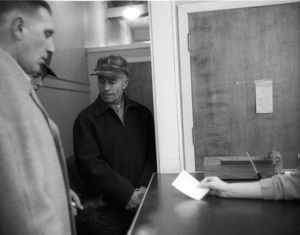 Ed Gein (1906-1984) and a police officer wait at the counter at the State Crime Lab where he was brought for lie detector and other tests or at the city jail where he was held overnight.