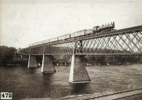 A day passenger train crossing the high North Western railroad bridge over the Chippewa River. Railroad tracks are along the shoreline in the foreground.