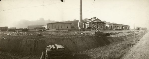 Shops under construction for the Chicago, St. Paul, Minneapolis, and Omaha railroad lines. Men are working with a horse-drawn wagon in the right foreground. A large smokestack is in the center among buildings, groups of men are working throughout the site, and railroad cars are along the tracks. Houses are along the top of a hill in the background.