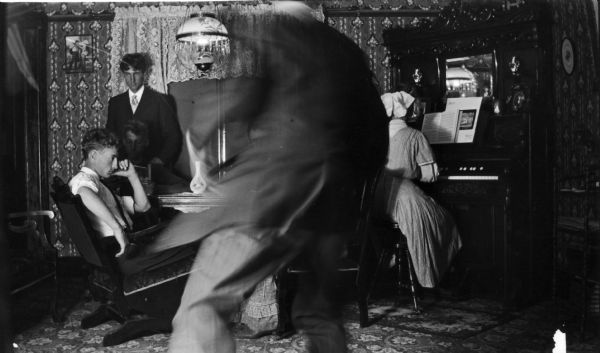 Living room scene in the Jacobson home, with a blurred image of a man running in front of the camera. A woman sits at a piano while three men are posed at a table.
