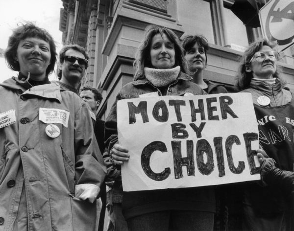 A group of pro-choice supporters stand together outdoors. One woman holds a sign that reads "Mother By Choice".