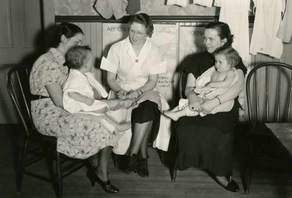 Nurse talking to two seated mothers; each with infant in their lap.
