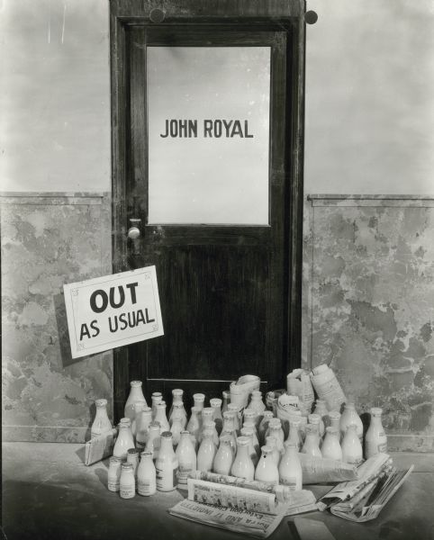View outside John Royal's office door. There are milk bottles and newspapers piled at the door and on it hangs a sign that reads "Out As Usual".