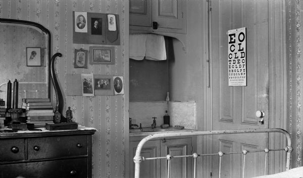 Interior view of a bedroom with an eye chart hanging on the door and a sink on the back wall.