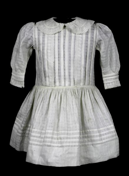 Ivory check-woven cotton; machine-sewn; slightly dropped waistline; straight, unfitted bodice; knee-length skirt, gathered into waistline; full puffed sleeves, gathered into tops of shoudlers as well as fitted cuffs; fitted neckline with large Peter Pan-style collar; 10 vertical pleats down front and back of bodice, 2 additional rows of machine-embroidered eyelet with scalloped edges added down front; machine-embroidered tape with ivory feather stitching covers waist seam; matching machine-embroidery edges collar as well as the machine-tape; cuffs finished by a row of tape, followed by a narrow pleat, another row of tape, and a row of eyelet; 4 horizontal pleats taken above hem of skirt, starting 3" up from the bottom; center back opening, open to waist seam, closes with 5 large off-white crochet-covered buttons.