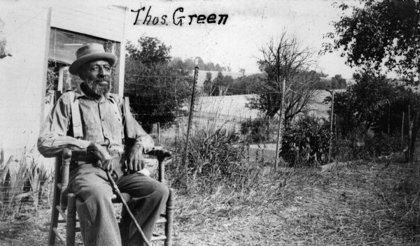 Portrait of Thomas Greene, seated outdoors in a chair, holding a cane. There is a fence and trees in the background.