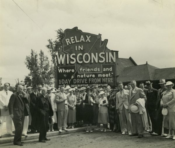 Group of resort and hotel owners gathered in front of a Wisconsin tourism sign that reads: "Relax In Wisconsin." There is a building in the background.