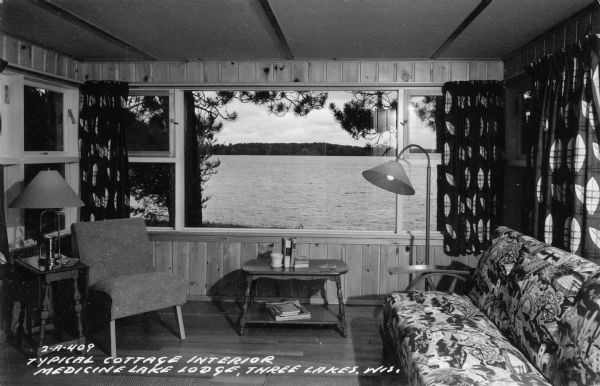 Cottage interior with a large window looking out on the lake. Caption reads: "Typical Cottage Interior, Medicine Lake Lodge, Three Lakes, Wis."