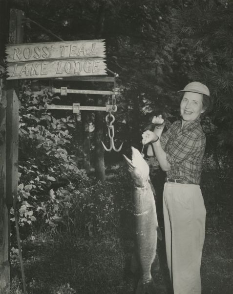 Virginia Hess posing with a muskellunge near the fish scale at Ross Teal Lake Lodge. Virginia Hess was a member of the Hess Oil Co. family, was from Chicago, and shared an apartment with Evelyn Ross Woods, younger sister of Virginia Ross.
