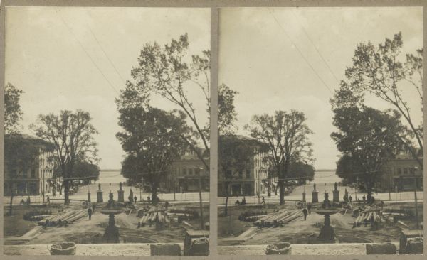 Stereograph view toward Lake Monona from the State Capitol during the construction of the fourth capitol building. Features the Capitol Square sidewalk (under construction), leading toward a water fountain. Lake Monona is on the horizon.
