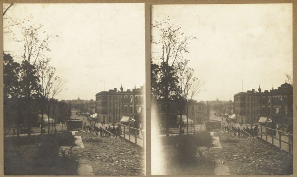 Stereograph view toward West Washington Avenue from the Wisconsin State Capitol during the construction of the fourth capitol building. Features the Capitol Square sidewalk staircase (under construction), with pedestrians in the street. A cable car is in the middle distance.