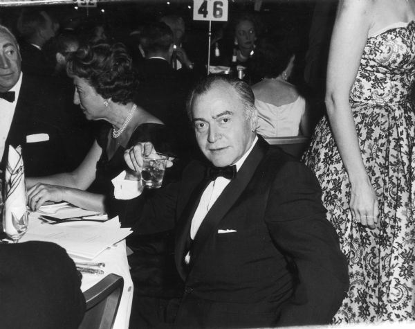 Kermit Bloomgarden is seated at a table at a formal event. He is wearing a tuxedo and holding a cocktail.