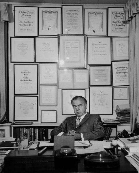Kermit Bloomgarden is seated at a desk with numerous awards on the wall behind him.