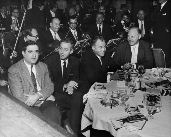 Kermit Bloomgarden seated at a table with three other men (he is the third from the left) at a formal event. Behind them sits a small orchestra.