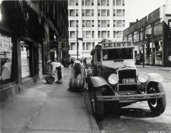Two men are rolling Leisy's beer barrels from an International truck on a downtown street. The truck was owned by Leisy Brewery.