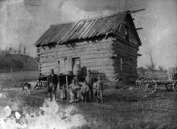 The family of Samuel Arms poses outdoors in front of their house (the front door faces east). From left to right are: Samuel Arms, Mamie, Mary Roberts Arms (holding Robert), William, Bernard (in rear), and Herman.