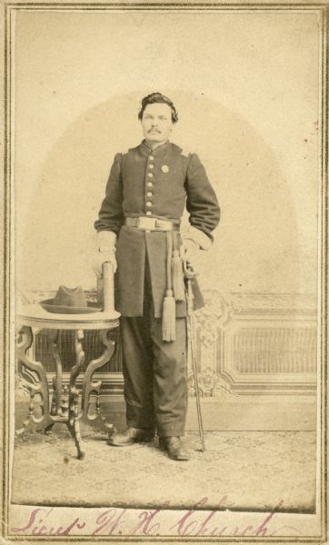 Full-length studio portrait of Lieutenant William H. Church standing next to a small table.