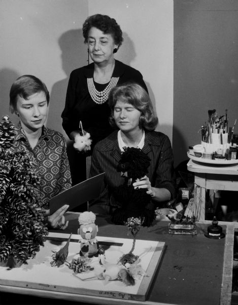 Aline W. Hazard stands behind two seated women who are making crafts.