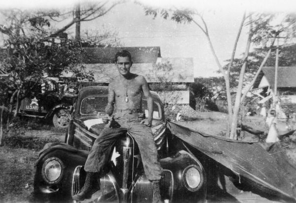Rod Serling sits shirtless on the hood of a car.