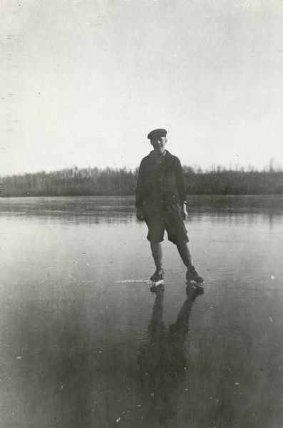 A young man, probably George Mattis, Jr., posing wearing ice skates on a frozen lake (possibly Knuteson Lake).