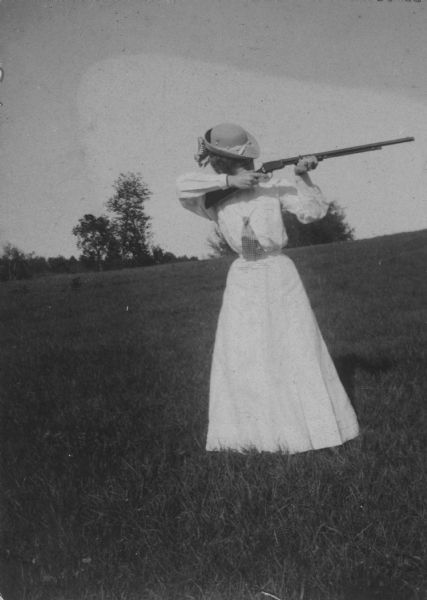 Woman in a dress and hat standing in a field and aiming a gun.