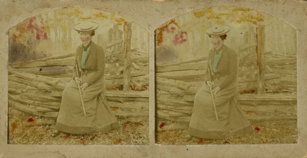 Hand-colored stereograph of Mattie Sprague Attoe Du Mond sitting on a rail fence and holding a .22 rifle near the fish pond off Pine River on John O. Evans' farm.