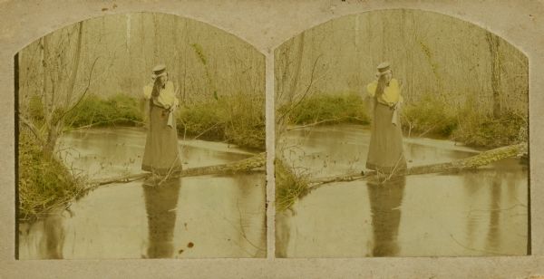 Hand-colored stereograph of Mattie Sprague Attoe Du Mond holding a .22 rifle and standing on a fallen tree limb over the fish pond off Pine River on the property of John O. Evans.