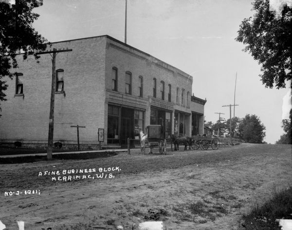 View down an unpaved Merrimac street with a brick building and horse-drawn carriages. The caption at bottom left reads: "A Fine Business Block."