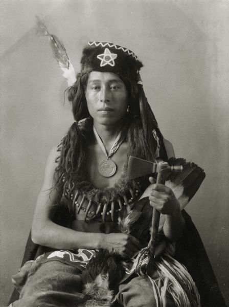 Studio portrait of Oto man, Cheedobau or Richard W. Shunatona. Part of Siouan (Sioux) and Otoe Tribes. He wears a medal and bear claw necklace, and holds an axe,