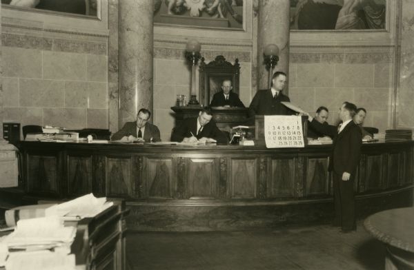 A messenger hands materials to Lawrence Larson, in the Senate Chamber of the Wisconsin State Capitol. From left to right are Jerry Czarnecki, Hanford Wesley, Lawrence Larson, John Danielson, and Paul Sawyer. Henry Gunderson sits behind them.