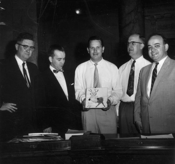 Senator Horace Wilke poses with four other men, one of whom is holding a drawing of a badger/deer creature.