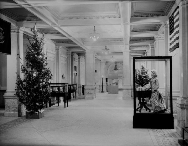 Christmas tree and costume exhibit in the first floor lobby of the State Historical Society building.  The view, based on identification in the floor mosaic is toward the south entrance.  Note: this view provides a good view of the lighting fixtures in the hallway.