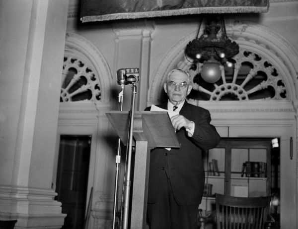 Wisconsin Supreme Court Justice, Marvin Rosenberry broadcasting over a WHA microphone located in the first floor lobby of the State Historical Society.  This photograph provides a good view of the original first floor lighting fixtures.