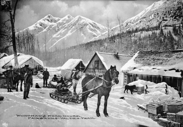 Exterior view of Wortman's Road House, on the Fairbanks-Valdez trail in Alaska, with horse-drawn sleds and people and dogs in front of a number of buildings. Mountains are in the background. Caption reads: "Wortman's Road House, Fairbanks-Valdez Trail."