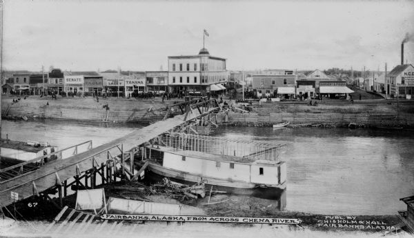 View of Fairbanks, Alaska across the Chena River. A wooden bridge crosses the river, and storefronts can be seen lining the city street. Caption reads: "Fairbanks, Alaska From Across Chena River."