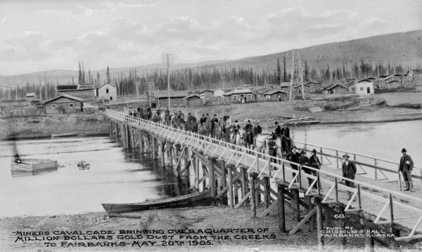 Elevated view of a group of miners on horseback crossing a bridge into Fairbanks, Alaska, May 20th, 1905. Houses are on the other side of the river. Caption reads: "Miners cavalcade bringing over a quarter of million dollars gold dust from the creeks to Fairbanks — May 20th 1905."