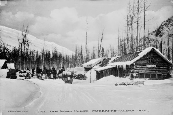 View across snow-covered road towards several men and horses in front of the Dam Road House on the Fairbanks-Valdez Trail. Caption reads: "The Dam Road House, Fairbanks-Valdez Trail."