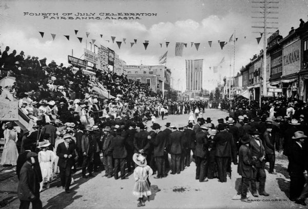 A large crowd of people gather for a 4th of July celebration. Sign on building on the right side of the street reads: "Tanana." A large American flag is hanging over the street in the background. Caption reads: "Fourth of July Celebration Fairbanks, Alaska."