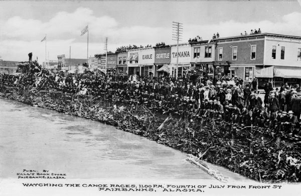 Elevated view across water towards a large crowd watching canoe races on the Fourth of July. Storefronts line the street behind the crowd. Business signs read: "Eagle", "Seattle" and "Tanana". Caption reads: "Watching The Canoe Races 11:00 P.M. Fourth of July from Front St Fairbanks, Alaska."