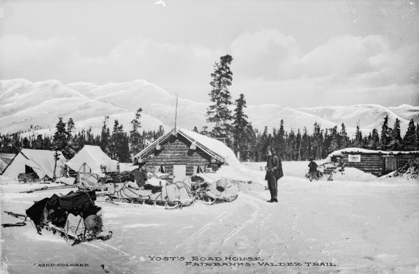 View of men and sleds outside Yost's Road House on the Valdez-Fairbanks Trail. Mountains can be seen in the background. Caption reads: "Yost's Road House, Fairbanks-Valdez Trail."