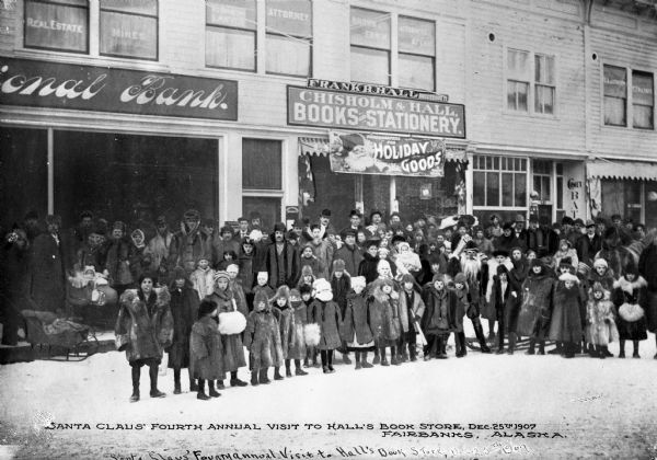 A crowd poses with Santa Claus in front of Hall's Book Store during a Christmas celebration. Business signs read, (partially obscured): "National Bank." Caption reads: "Santa Claus' Fourth Annual Visit To Hall's Book Store, Dec. 25th, 1907, Fairbanks, Alaska."
