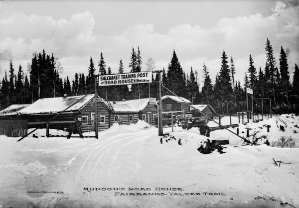 Exterior view of the Salchaket Trading Post and Munson's Road House on the Valdez-Fairbanks Trail. Log buildings are along a snowy path. Caption reads: "Munson's Road House. Fairbanks-Valdez Trail."