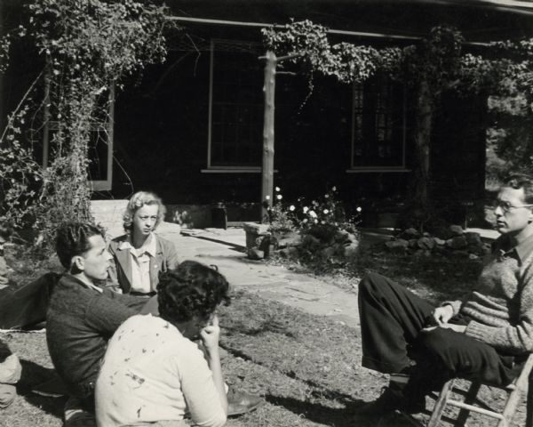 Four people have an informal meeting outdoors at Highlander School. Three of them are seated on the grass and one is seated on a chair. Myles Horton on the far right.